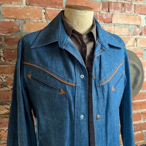 K&ouml;nnte beinhalten: Eine blaue Jeansjacke mit braunen Zierleisten und Taschen. Die Jacke hat eine Knopfleiste vorne und einen breiten Kragen. Darunter ist ein braunes Hemd mit Kragen zu sehen. Die Jacke wird auf einer Schaufensterpuppe vor einer Backsteinmauer pr&auml;sentiert.