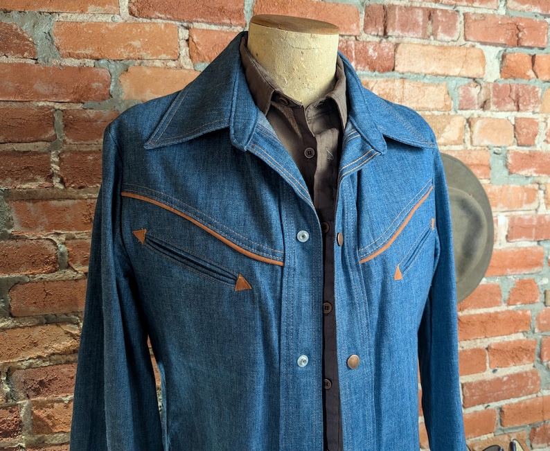 K&ouml;nnte beinhalten: Eine blaue Jeansjacke mit braunen Zierleisten und Taschen. Die Jacke hat eine Knopfleiste vorne und einen breiten Kragen. Darunter ist ein braunes Hemd mit Kragen zu sehen. Die Jacke wird auf einer Schaufensterpuppe vor einer Backsteinmauer pr&auml;sentiert.