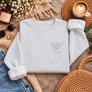 Könnte beinhalten: Ein hellgraues Sweatshirt mit Rundhalsausschnitt, das auf der linken Brustseite eine kleine, detaillierte violette Strichzeichnung einer Biene sowie den darunter gedruckten Text "I'm a bee" in einer schlichten Schriftart zeigt.