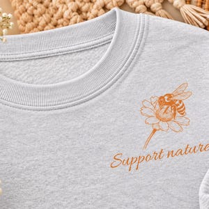 Könnte beinhalten: Ein hellgraues Sweatshirt mit Rundhalsausschnitt und einer orangefarbenen Grafik einer Biene auf einem Gänseblümchen. Unter der Blumenillustration ist der Text "Support nature." in passender orangefarbener Schreibschrift aufgedruckt.