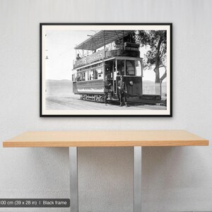 May include: A black and white framed wall art print featuring a vintage double-decker tram with the text 'LEGGO'S PICKLES' on the side. Two individuals stand in front of the tram. The frame measures 100 by 70 cm and is black in colour.