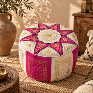 May include: A round ottoman featuring a vibrant fuchsia and cream color palette. The top surface displays a central circular gold sunburst motif surrounded by alternating fuchsia and cream triangular panels with gold floral accents and black trim.