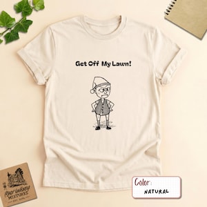 Może przedstawiać: T-shirt w naturalnym kolorze z czarną ilustracją liniową przedstawiającą osobę w czapce i kamizelce, z rękami na biodrach. Nad postacią znajduje się napis Get Off My Lawn! wykonany pogrubioną, czarną, zaokrągloną czcionką.