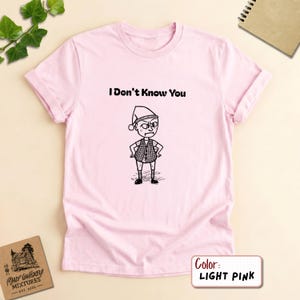 May include: A light pink short-sleeved t-shirt featuring a black line drawing of a character wearing a hat and a vest with the text "I Don't Know You" printed in black above the illustration. The shirt has a crew neckline and rolled sleeves.
