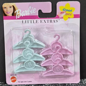 May include: A Barbie Little Extras blister pack containing six plastic doll clothes hangers. Three hangers are light blue and three are light pink. Each hanger features a circular center detail with the letter B embossed on the front surface.
