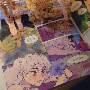 Pode incluir: Uma p&aacute;gina de banda desenhada com ilustra&ccedil;&otilde;es coloridas. A obra apresenta uma personagem com cabelo branco segurando uma flor azul. Bal&otilde;es de texto incluem frases como "Eu posso fazer algo sobre isso" e "Est&aacute; feito!" Flores secas no fundo.