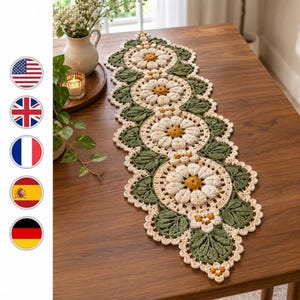 May include: A long, rectangular crochet table runner featuring a repeating pattern of large, cream-colored daisy motifs with golden-yellow centers, interspersed with textured, dark green leaf designs and a scalloped, cream-colored border edge.