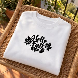 Könnte beinhalten: Ein weißes Sweatshirt mit Rundhalsausschnitt und einem schwarzen Grafikdruck auf der Brust. Das Design zeigt den Text "Hello Fall" in einer Schreibschrift, umgeben von zwei Ahornblättern, einem Eichenblatt und einer Eichel in Schwarz.