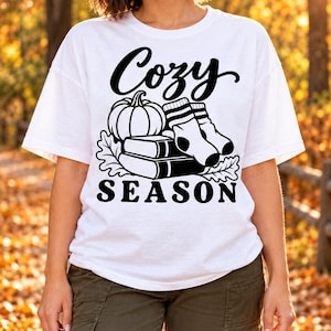Könnte beinhalten: Ein weißes Kurzarm-T-Shirt mit einem schwarzen Grafikdruck, der einen Kürbis, ein Paar gestreifte Socken und zwei gestapelte Bücher zeigt. Der Text "Cozy" steht in Schreibschrift darüber und das Wort "SEASON" darunter.