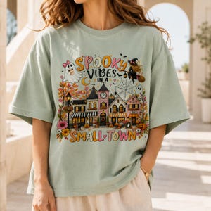 May include: A sage green t-shirt featuring a graphic print of a small town with a clock tower, pumpkins, and autumn foliage. The text reads "SPOOKY VIBES IN A SMALL TOWN" with illustrations of a ghost, a witch on a broomstick, and a spider web.