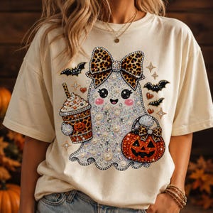 May include: A cream-colored t-shirt featuring a graphic of a ghost adorned with rhinestones and pearls. The ghost wears a leopard print bow, holds a leopard print coffee cup with a pumpkin, and carries a jack-o'-lantern bucket with bats nearby.