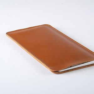 May include: A rectangular tan leather sleeve case with rounded corners and visible stitching along the edges. The sleeve has a smooth, matte finish and contains a thin, white electronic device partially inserted into the open top end.