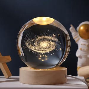 May include: A clear glass sphere lamp featuring an internal laser-etched golden spiral galaxy design with small stars. The sphere rests on a cylindrical light-colored wooden base with an attached white power cord extending to the right side.