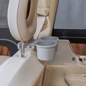 Puede incluir: Un soporte doble para vasos de color gris claro montado en los postes metálicos de un reposacabezas de coche beige. El soporte presenta una superficie exterior texturizada y acanalada con dos aberturas circulares para recipientes.