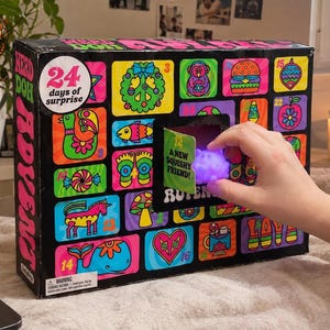 May include: A black rectangular advent calendar box with 24 numbered windows featuring colorful neon-style illustrations of animals, food, and symbols. One open window reveals a small purple squishy toy with the text A NEW SQUISHY FRIEND.