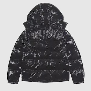 Könnte beinhalten: Eine schwarze Daunenjacke mit glänzender, reflektierender Oberfläche und Kapuze. Auf der Rückseite ist das Wort TRAPSTAR in einer gotischen Schriftart im oberen Bereich eingestickt. Die Jacke hat horizontale Steppnähte und lange Ärmel.