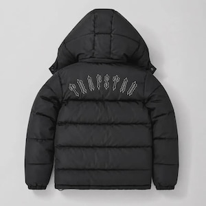 Könnte beinhalten: Eine schwarze Daunenjacke mit Kapuze und horizontalen Steppnähten. Das Wort TRAPSTAR ist in einer weißen Schriftart im gotischen Stil auf den oberen Rückenbereich des Kleidungsstücks gestickt, direkt unter dem Ansatz der Kapuze.