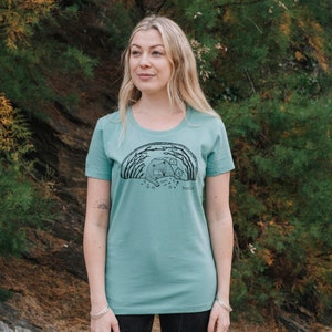 May include: A teal blue t-shirt with a black and white illustration of a bear and cubs in a forest. The text "boo-cle" is printed below the illustration.