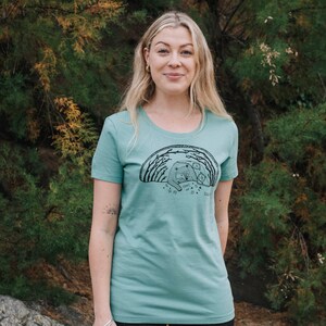May include: A teal blue t-shirt with a black graphic of a bear and cubs in a forest. The graphic includes the text "Grow Wild, Be Kind, Do Good".