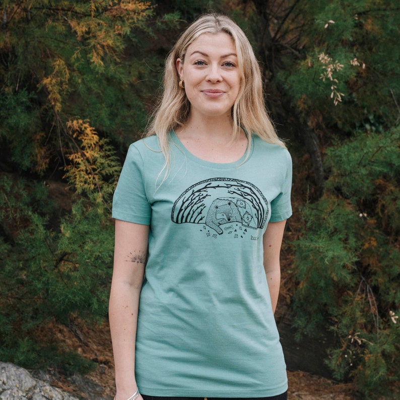 May include: A teal blue t-shirt with a black graphic of a bear and cubs in a forest. The graphic includes the text "Grow Wild, Be Kind, Do Good".