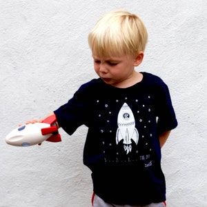 May include: A young child wearing a navy blue t-shirt with a white rocket ship graphic and the text "THE 3, 2, 1...  THE  BOOTY" printed on the bottom. The child is holding a white toy rocket ship with red accents.