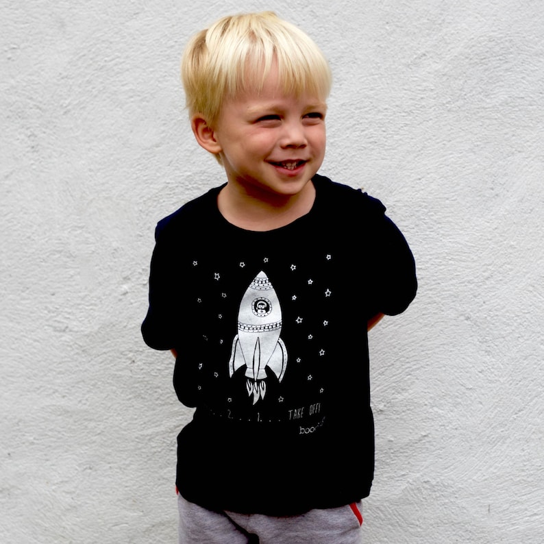 May include: A young boy wearing a black t-shirt with a white rocket ship graphic and the text "Take off, boo!" printed on it.