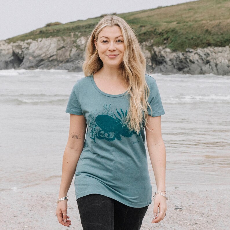 May include: A teal blue t-shirt with a dark blue octopus graphic print. The octopus is surrounded by seaweed and coral. The shirt has a scoop neck and short sleeves.