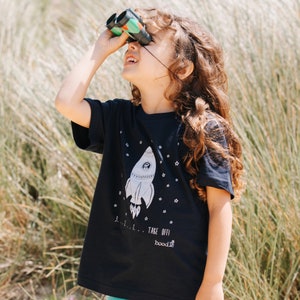 May include: A black t-shirt with a white rocket ship graphic and the text "3...2...1... TAKE OFF! boodie" printed on the front. A child is wearing the shirt and looking through binoculars.