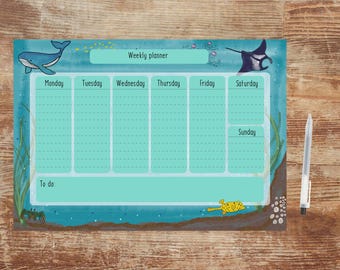 May include: A weekly planner with an ocean theme. The planner features illustrations of a whale, stingray, and other sea creatures. The text "Weekly planner" is at the top, with days of the week listed below. A pen rests on the right side.