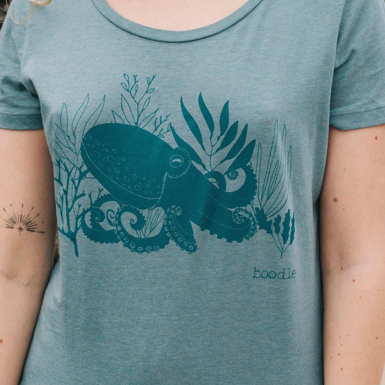 May include: A teal blue t-shirt with a graphic print of an octopus surrounded by seaweed. The text "boodle" is printed on the bottom right corner of the shirt.