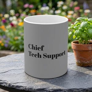 Op de afbeelding: Een matte lichtgrijze keramische mok met de zwarte tekst Chief Tech Support op de voorzijde. De mok heeft een cilindrische vorm met een witte binnenrand en staat rechtop op een donker, getextureerd oppervlak.