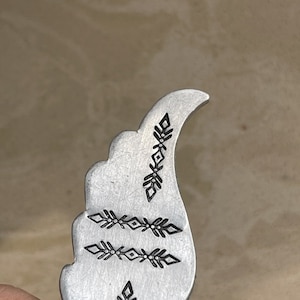 May include: A silver-coloured metal pendant with a curved, wing-like shape. The pendant features three rows of black, stylised arrow and leaf designs. The metal has a brushed texture, and the overall design is minimalist and geometric.