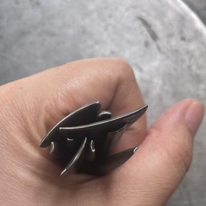 May include: A silver metal ring with a unique, abstract design. The ring is made of multiple pieces of metal that are stacked and intertwined.