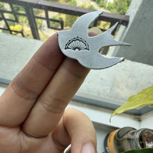 May include: A silver-colored metal bird-shaped pendant. The pendant features a detailed design of a stylized sun or flower on its body. The bird's wings are outstretched, and the pendant is held up in front of a window.