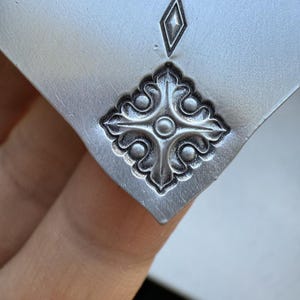May include: A silver metal pendant with a decorative design featuring a cross with four rounded points and a circle in the center. The cross is surrounded by four stylized leaves.