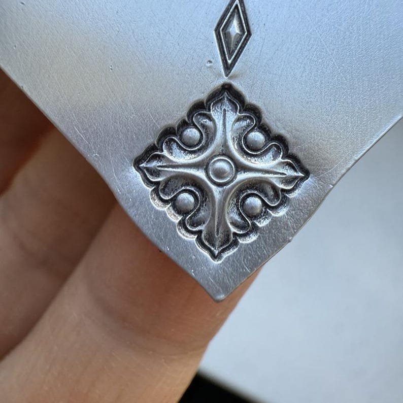 May include: A silver metal pendant with a decorative design featuring a cross with four rounded points and a circle in the center. The cross is surrounded by four stylized leaves.