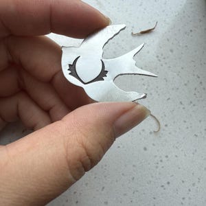 May include: A silver-colored metal bird-shaped pendant. The pendant has a cut-out design in the center and is held by a person's hand. The bird's wings are spread, and the design is detailed.