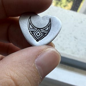 May include: A heart-shaped, silver-toned pendant with an etched tribal design. The design features a crescent shape above a geometric pattern with a central square. The pendant is held between fingers.