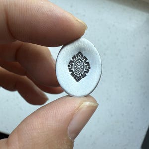 May include: A small, oval-shaped, silver-colored pendant with a stamped black geometric design. The design features a central cross-like shape surrounded by a patterned border. The pendant is held between two fingers.