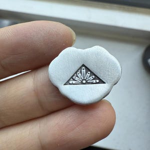 May include: A small, silver-colored, flower-shaped pendant. The pendant features a triangle with a floral design inside. The pendant is held in a person's hand, and the background is out of focus.