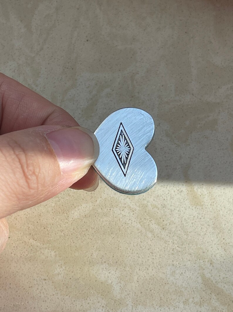 May include: A heart-shaped silver-toned metal charm with a brushed finish. The center of the charm features a black engraved diamond-shaped border containing a stylized sunburst or floral pattern with radiating lines.