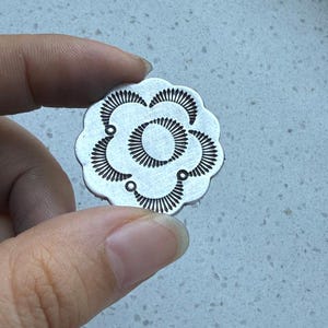 May include: A circular silver-toned metal disc featuring a scalloped edge and a stamped floral pattern. The design consists of a central oval surrounded by five petal-like shapes, each filled with fine radiating lines and small circular accents.