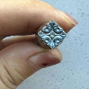 May include: A silver metal stamp with a square design featuring a decorative floral pattern. The stamp has a raised center circle.