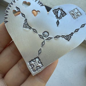 May include: A silver heart-shaped metal blank with a decorative design of diamonds, hearts, and floral patterns.