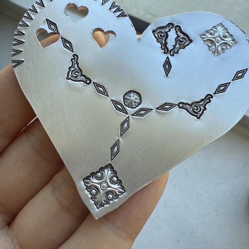 May include: A silver heart-shaped metal blank with a decorative design of diamonds, hearts, and floral patterns.