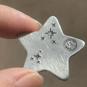 May include: A star-shaped silver-toned metal charm with a brushed finish. The surface features stamped designs of two small star clusters and a circular moon icon. The metal has visible fine scratches across its flat, five-pointed surface.