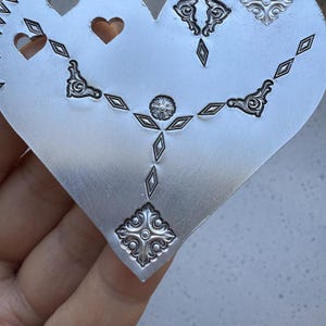 May include: A silver heart-shaped metal sheet with a decorative design featuring a repeating pattern of diamonds, hearts, and floral motifs.
