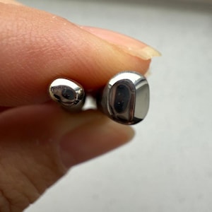 May include: Close-up of two small, shiny, silver-coloured metal objects. One is oval-shaped, and the other is a rounded rectangle. The objects are held between fingers, suggesting they are small accessories or components.