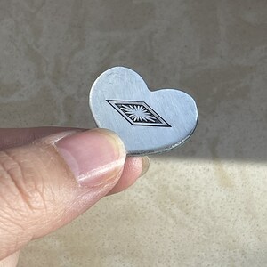 May include: A small, heart-shaped silver-toned metal charm featuring a centered engraved diamond-shaped outline containing a detailed sunburst or starburst pattern. The surface of the metal has a smooth, matte finish with clean, defined edges.