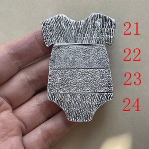 Puede incluir: Un dije de metal plateado con forma de body para bebé, que presenta cuatro bandas horizontales distintas con patrones texturizados. La superficie muestra variados detalles en relieve, y los números 21, 22, 23 y 24 están impresos en rojo a la derecha.
